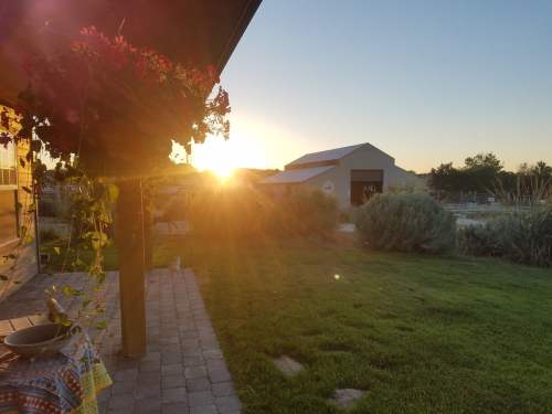 Sunset view from a porch with flowers, greenery, and a barn in the background, casting warm golden light.