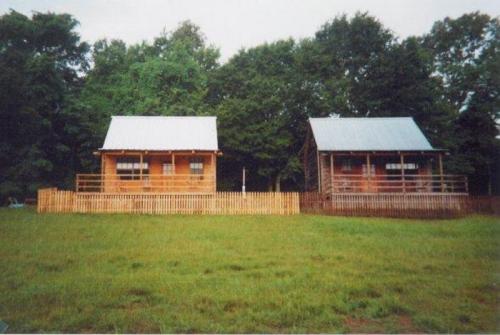 Two wooden cabins with metal roofs, surrounded by green grass and trees, separated by a wooden fence.