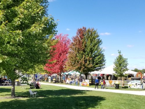 A park scene with colorful autumn trees, people walking, and tents set up for an outdoor event.