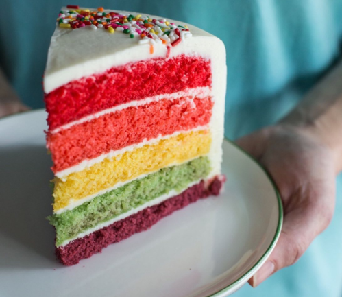 A slice of colorful layered cake with red, orange, yellow, green, and purple layers, topped with sprinkles.