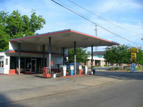 A small gas station with a red and white canopy, vintage pumps, and a sign displaying fuel prices on a sunny day.