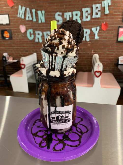 A tall dessert in a jar with ice cream, chocolate sauce, whipped cream, an Oreo, and a colorful straw on a purple plate.