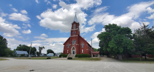A red brick church with a tall steeple, surrounded by trees and a gravel path under a partly cloudy sky.