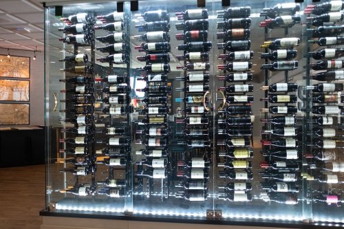 A glass wine display featuring numerous bottles arranged neatly on shelves, illuminated from below.