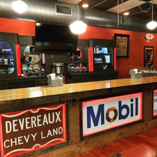 A retro diner interior featuring a wooden counter with signs for Devereaux Chevy Land and Mobil, and vintage decor.