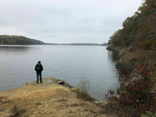 A person stands on a rocky shore, gazing out at a calm lake surrounded by trees in autumn colors under a cloudy sky.