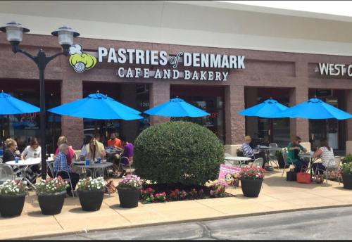 Outdoor seating at Pastries of Denmark café and bakery, with blue umbrellas and flower planters, bustling with patrons.