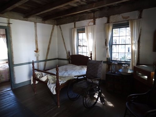 A rustic bedroom featuring a wooden bed, a wheelchair, and large windows with sheer curtains.