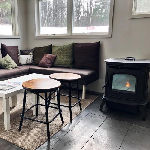 Cozy living room with a sofa, two wooden stools, a coffee table, and a wood stove, surrounded by large windows.