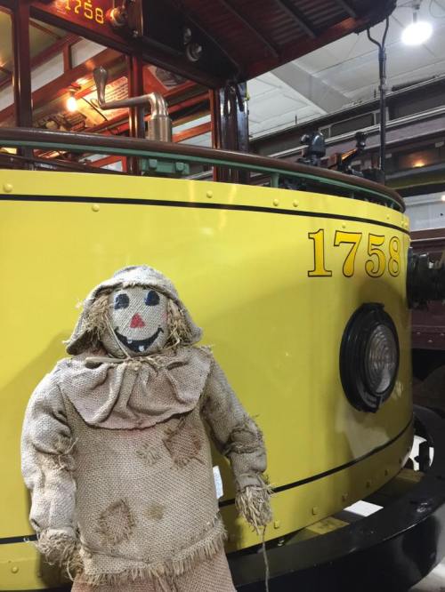 A scarecrow stands next to a vintage yellow train car marked "1758" in a museum setting.