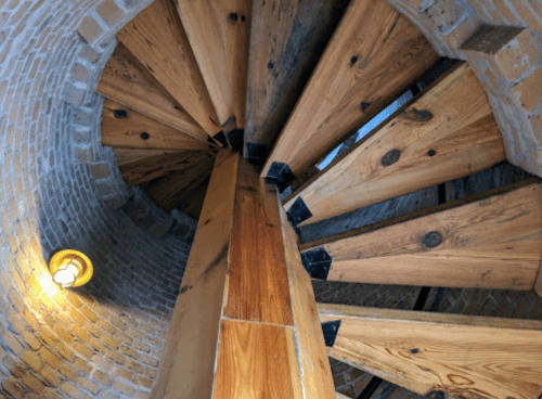 A spiral wooden staircase viewed from above, featuring a brick wall and a warm light fixture.
