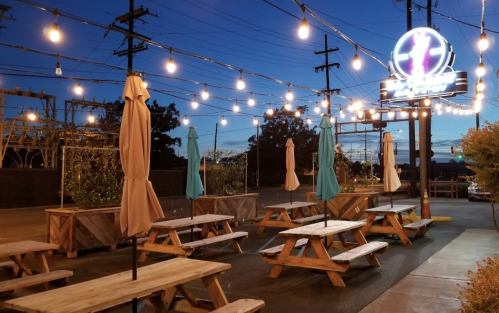 Outdoor dining area with wooden picnic tables, umbrellas, and string lights, set against a twilight sky.