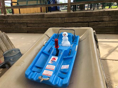 A blue sled on a slide with a small snowman sitting on it, surrounded by a playground setting.