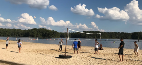 A group of people playing beach volleyball on a sandy shore by a lake under a blue sky with fluffy clouds.