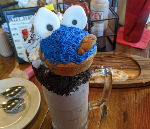 A whimsical milkshake topped with a cookie monster donut, whipped cream, and crushed cookies in a glass mug.