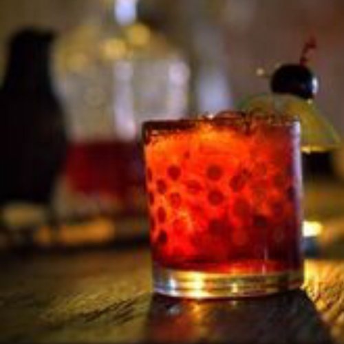 A close-up of a vibrant red cocktail in a glass, garnished with a cherry and a slice of lemon, on a wooden table.