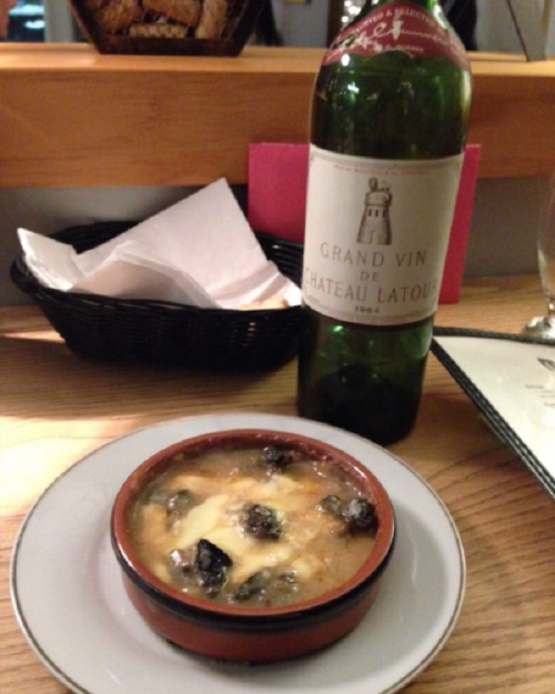 A small dish of baked cheese with mushrooms beside a bottle of red wine on a wooden table.