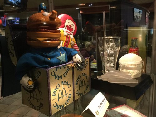 A display featuring a plush burger character, Ronald McDonald, and vintage McDonald's memorabilia in a glass case.