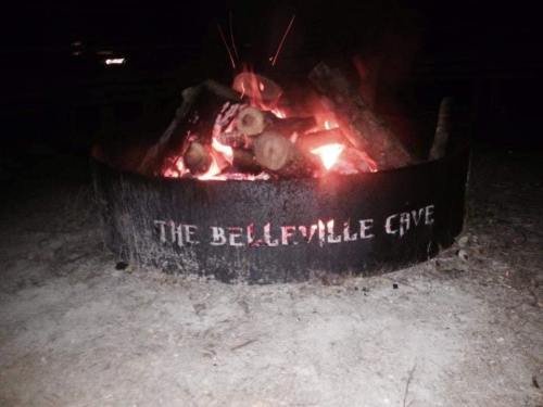 A campfire in a circular fire pit with the words "THE BELLEVILLE CAVE" visible on the side, surrounded by dark surroundings.