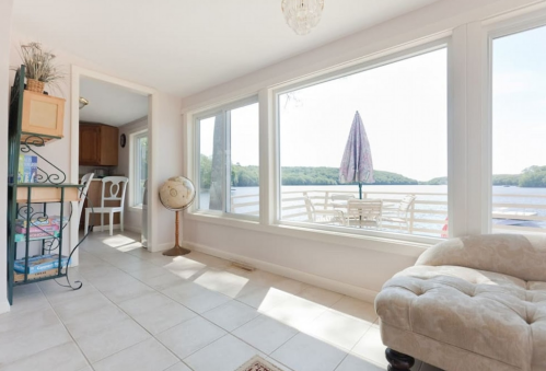 Bright, airy room with large windows overlooking a lake, featuring a cozy chair and a dining area.