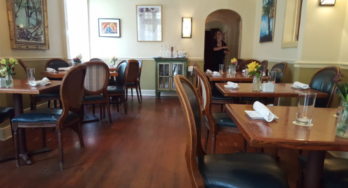 A cozy restaurant interior with wooden tables, chairs, and flower centerpieces, featuring a staff member in the background.