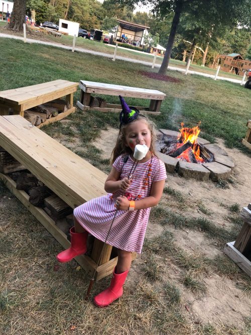 A girl in a striped dress and pink boots holds a marshmallow on a stick, sitting by a campfire with a party hat on.