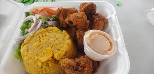 A takeout container with fried chicken, yellow rice, salad, and a small cup of dipping sauce.