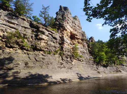 A rocky cliff rises above a calm river, surrounded by trees under a clear blue sky.