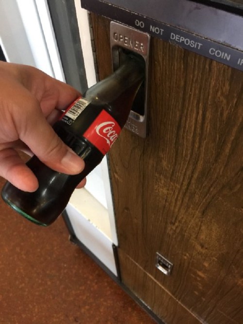 A hand holding a glass Coca-Cola bottle, inserting it into a bottle opener on a vending machine.