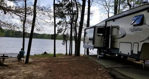 A camper parked by a serene lake, surrounded by trees, with a person sitting at a picnic table nearby.