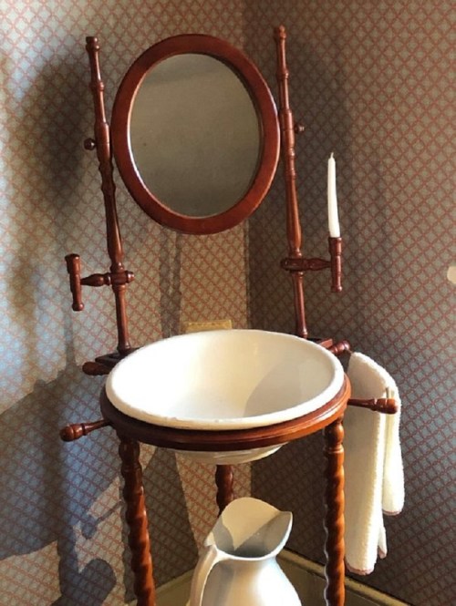 Antique washstand with a round mirror, white basin, pitcher, and towel hanging on a patterned wallpaper background.