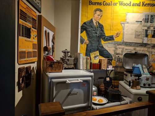 A vintage kitchen display featuring old appliances, utensils, and a large advertisement poster in the background.