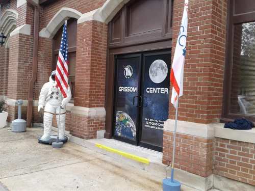 Entrance to the Grissom Center, featuring an astronaut statue and flags, with a moon and Earth graphic on the door.