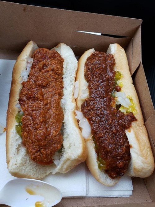 Two hot dogs in a cardboard container, topped with chili, onions, and pickles.