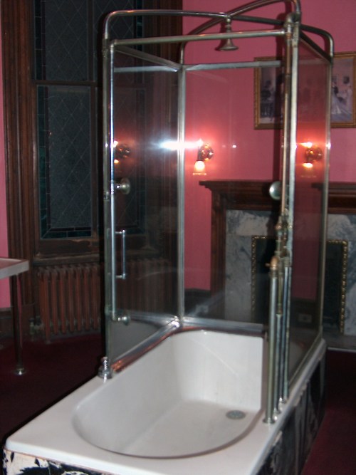 A vintage glass shower enclosure with a white bathtub in a pink-walled bathroom, featuring ornate fixtures.