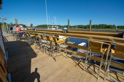 A waterfront deck with bar stools and tables, overlooking a marina with boats and clear blue skies.