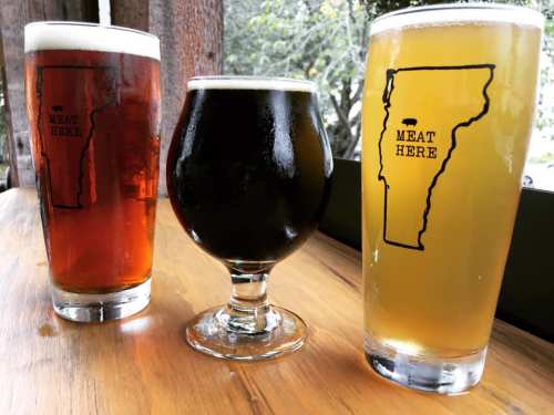 Three glasses of beer on a wooden table, featuring different colors and a "Meat Here" logo shaped like Vermont.