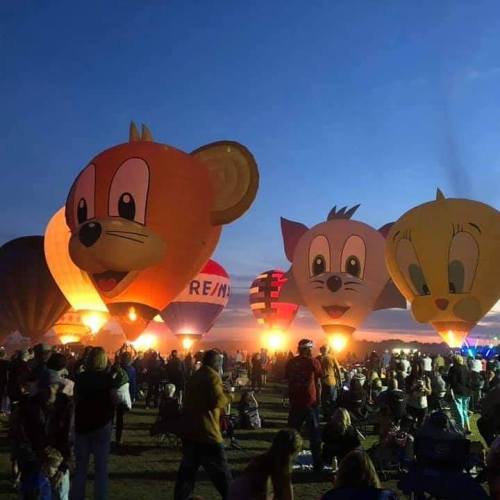 Colorful hot air balloons shaped like cartoon characters at sunset, with a crowd enjoying the event below.