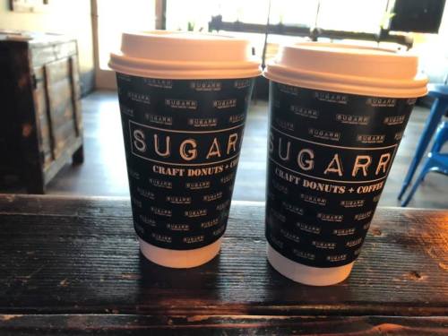 Two coffee cups with a black and white design, labeled "SUGAR" and "CRAFT DONUTS + COFFEE," on a wooden table.