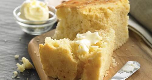 A slice of freshly baked cornbread with melted butter on a wooden cutting board, accompanied by a small bowl of butter.