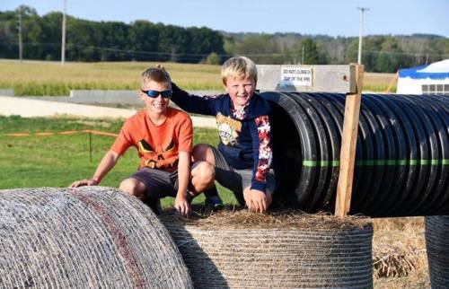Two boys playfully pose on top of hay bales and large black tubes in a sunny outdoor setting.