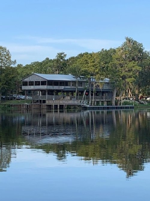 A wooden building with a deck sits by a calm lake, surrounded by trees and reflecting in the water.