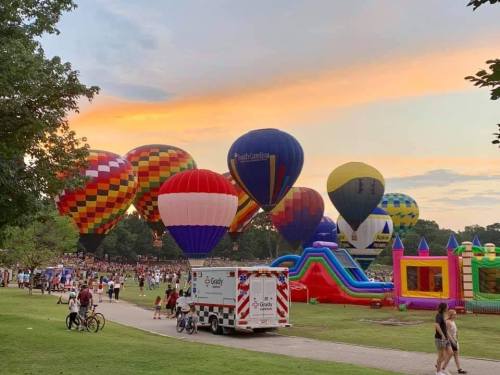 Colorful hot air balloons fill the sky at sunset, with a crowd and inflatable attractions in a park setting.