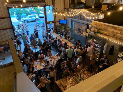 A lively indoor gathering with people socializing at tables, string lights overhead, and an open area visible outside.