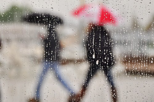 Two people walk with umbrellas in the rain, viewed through a window with droplets of water.