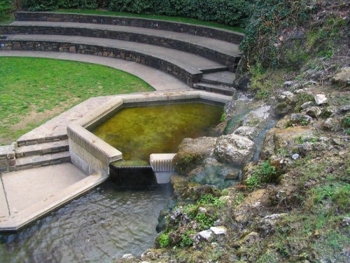 A serene outdoor space featuring a small pond, stone seating, and a gentle waterfall surrounded by greenery.