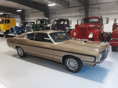 A classic gold car parked in a showroom, with vintage trucks displayed in the background.