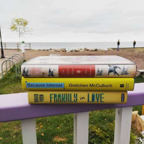 A stack of three books on a railing, with a beach and cloudy sky in the background.