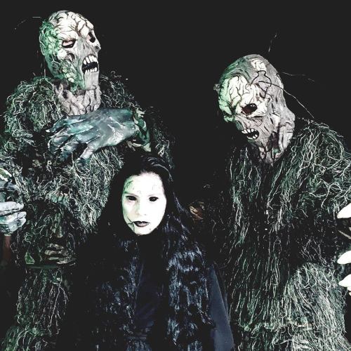 A girl with long black hair stands between two eerie, moss-covered figures with grotesque faces in a dark setting.