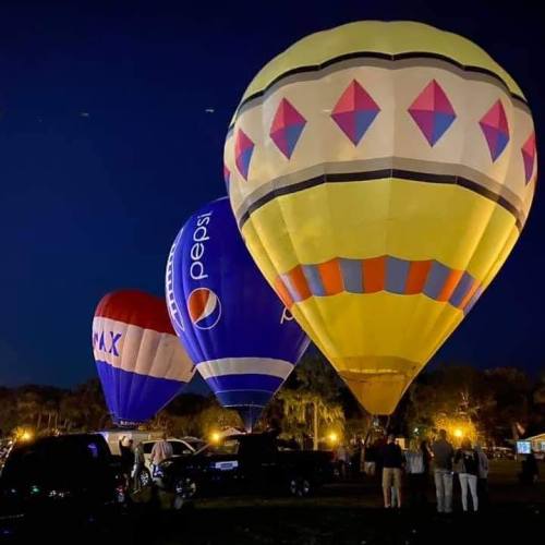 Three colorful hot air balloons illuminated at night, with people and vehicles nearby under a dark sky.